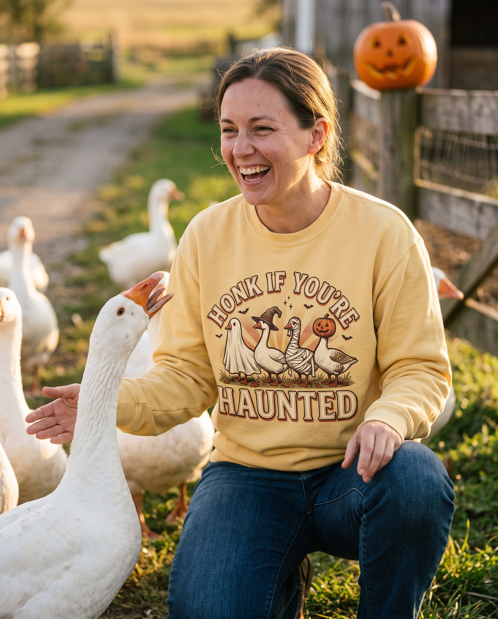 Woman in a Butter Funny Halloween Goose crewneck sweatshirt crouched in a farmyard beside a flock of white geese, one hand out as a goose honks at her, laughing.