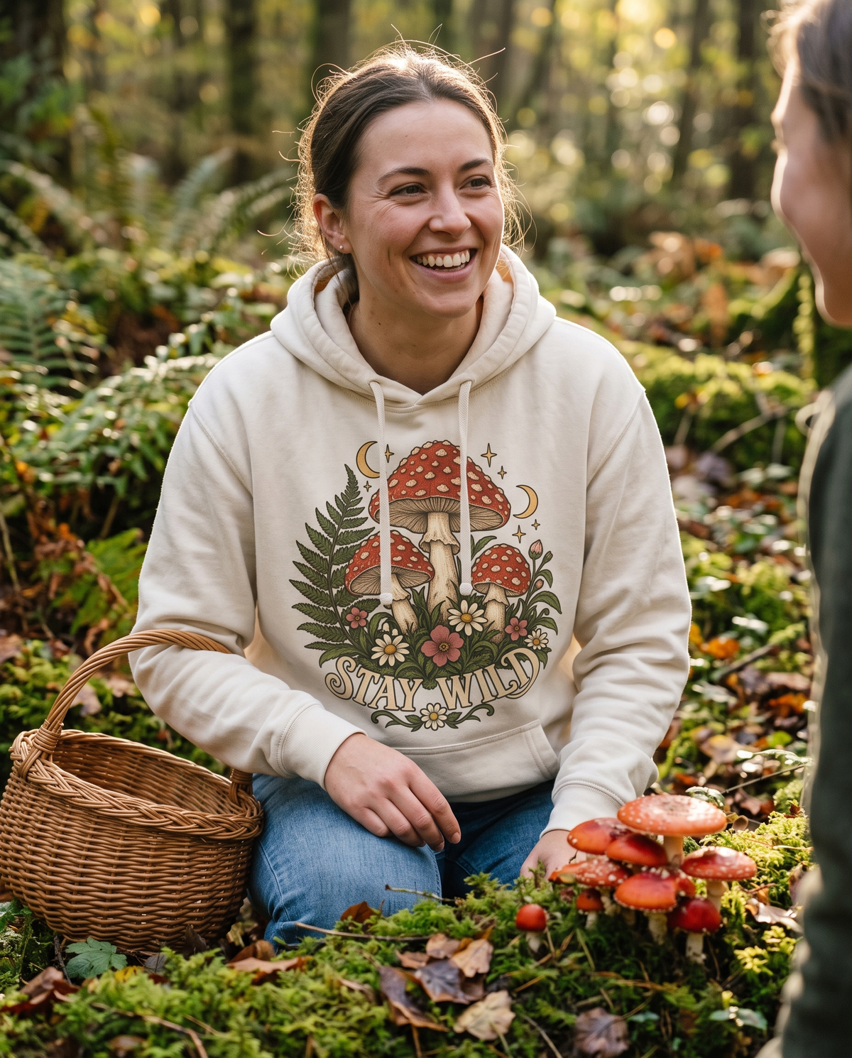 Woman in a White Stay Wild mushroom hoodie crouched on a mossy forest floor beside red capped toadstools with a wicker foraging basket