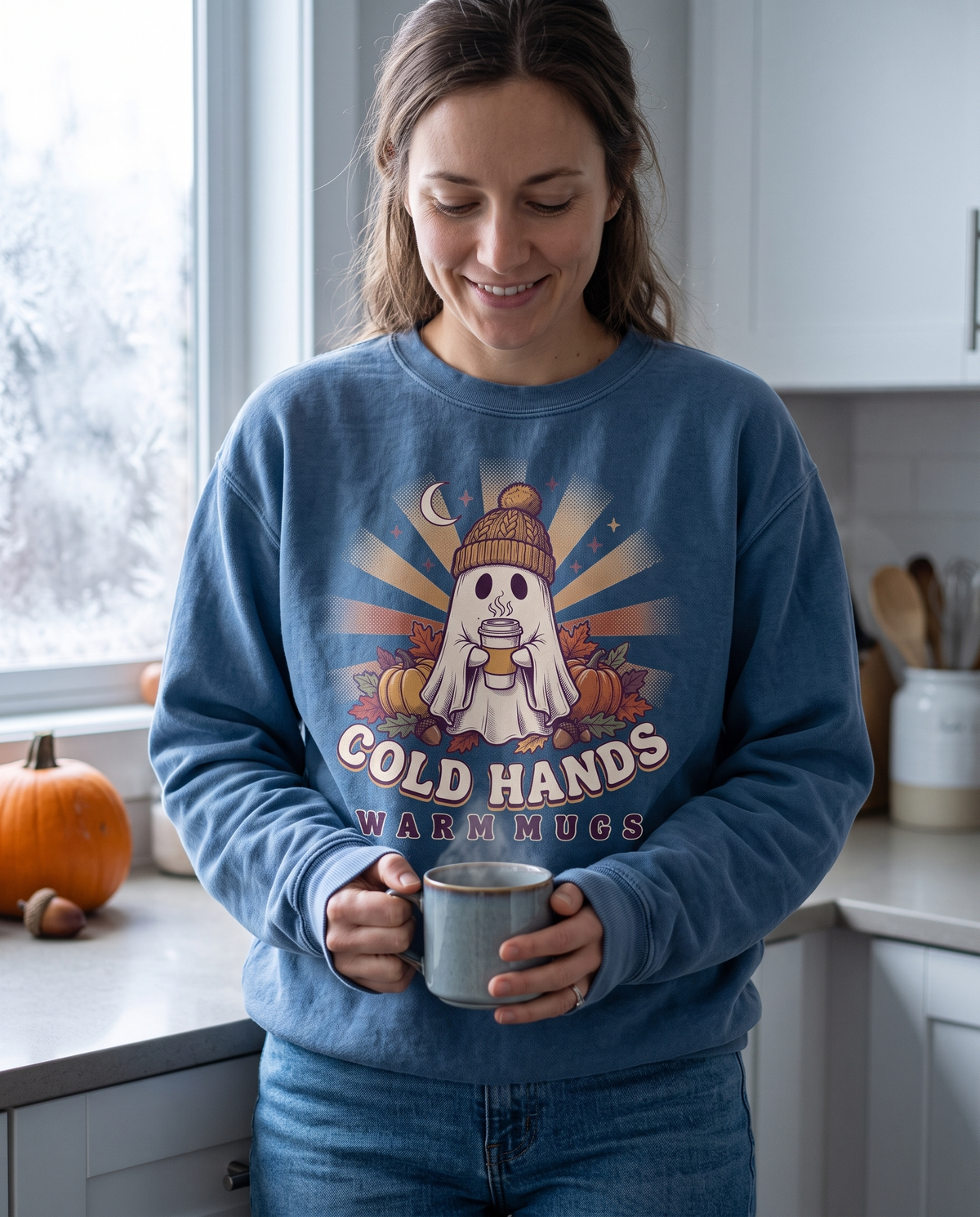 Woman in a Denim Cozy Ghost Cold Hands Warm Mugs crewneck sweatshirt holding a mug at a kitchen window with a pumpkin on the sill