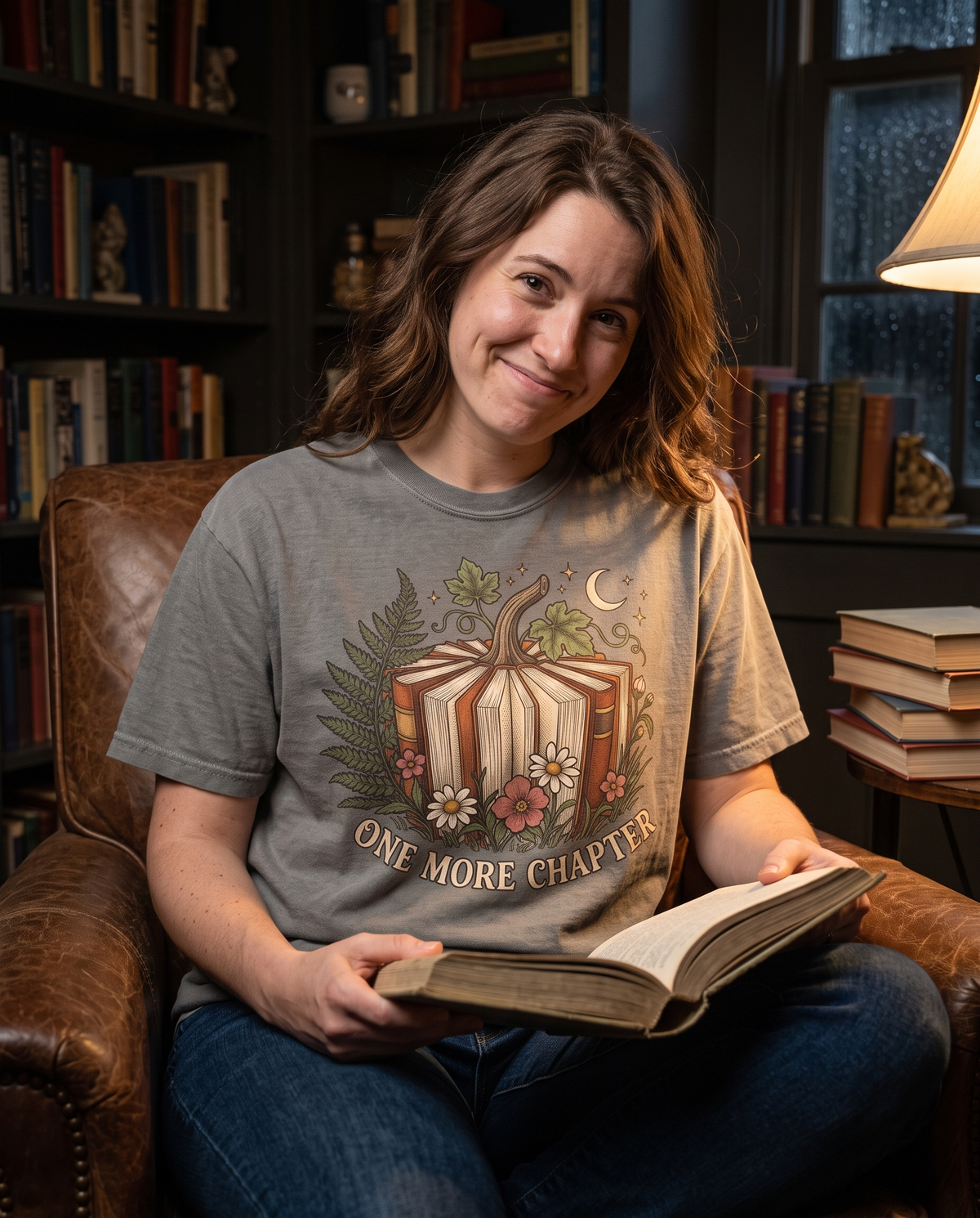Woman in a Granite Comfort Colors tee with the Book Lover One More Chapter print, curled in an armchair holding an open hardback under a reading lamp.