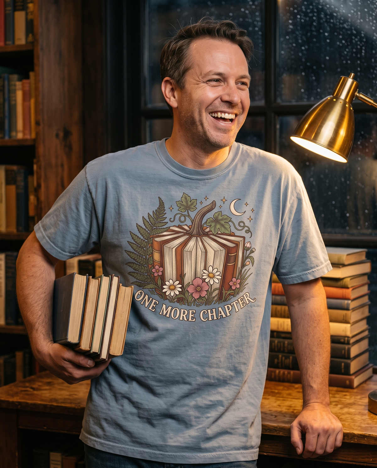 Man in a Blue Jean Comfort Colors tee with the Book Lover One More Chapter print, leaning on a bookshop counter with a stack of hardbacks under one arm.