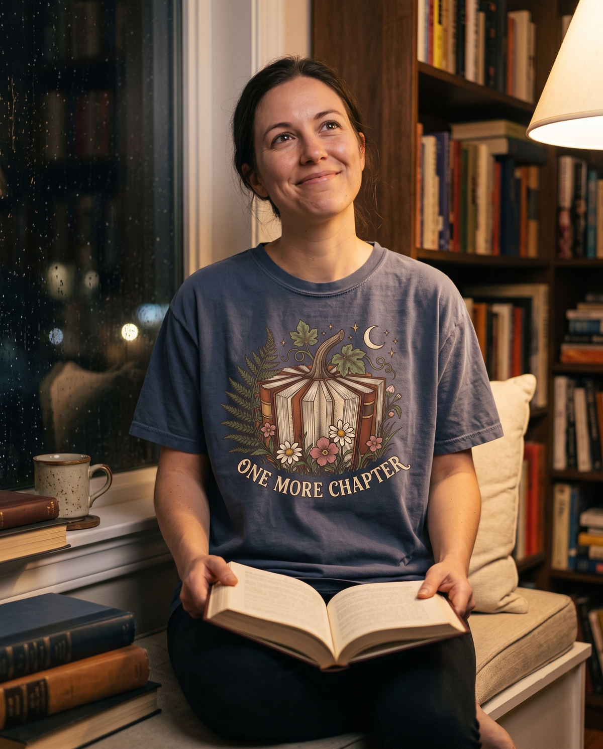 Woman in a Denim Comfort Colors tee with the Book Lover One More Chapter print, sitting on a window seat with a hardback open on her knee.