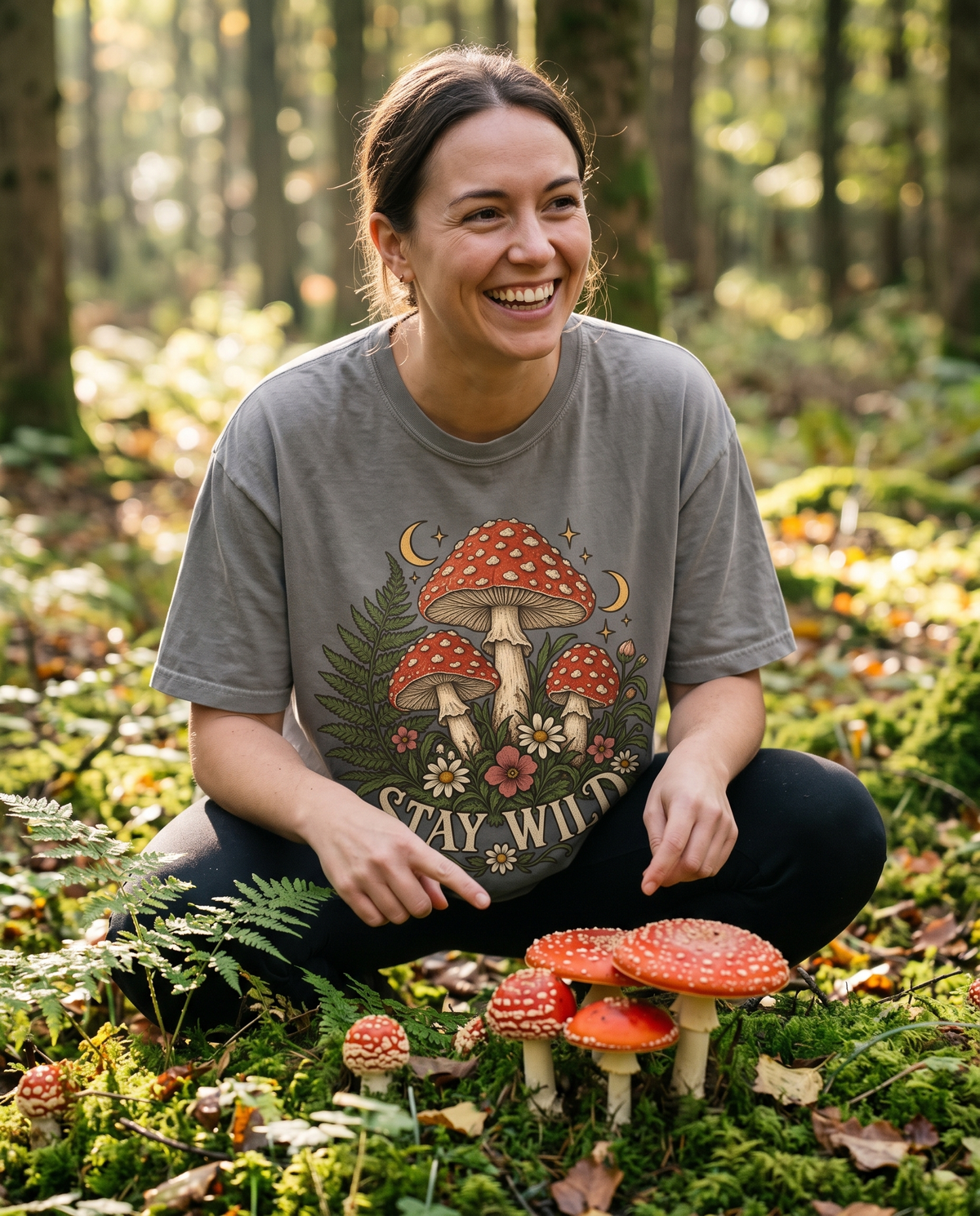 Woman in a Granite Comfort Colors tee with the Mushroom Vintage Botanical Toadstool print, crouched on a mossy forest floor beside red-capped toadstools.