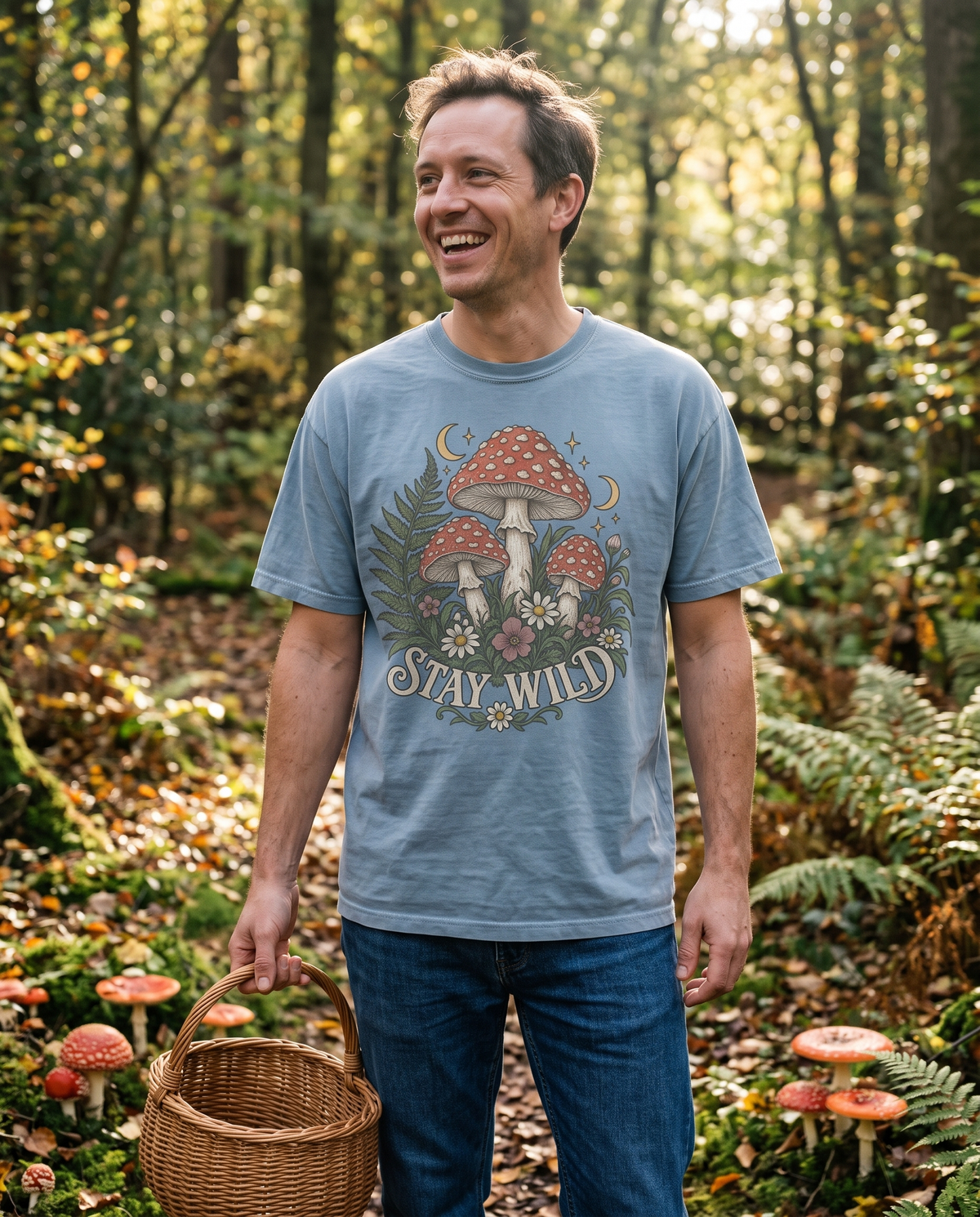 Man in a Blue Jean Comfort Colors tee with the Mushroom Vintage Botanical Toadstool print, standing on a woodland trail holding a wicker basket with toadstools at his feet.