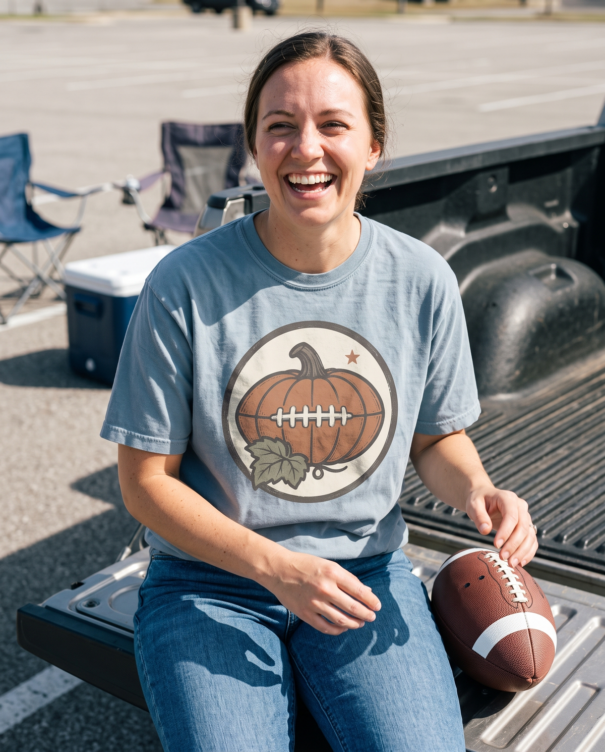 Woman in a Blue Jean Comfort Colors tee with the Pumpkin Football Vintage Fall Saturday Tailgate print, sitting on an open truck tailgate laughing with a football beside her.
