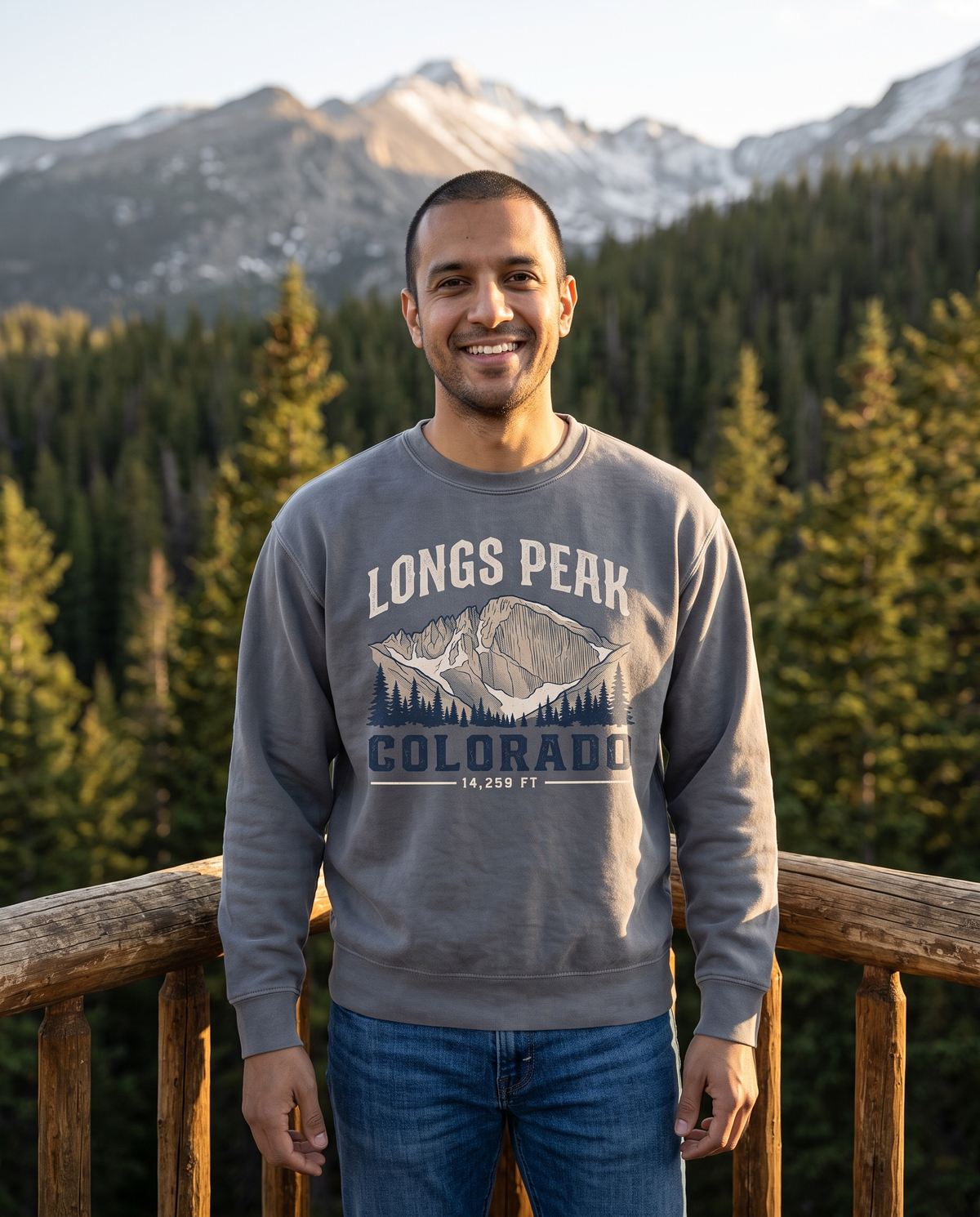 A man stands at a log deck railing with a forested valley and snow-streaked peaks behind him.