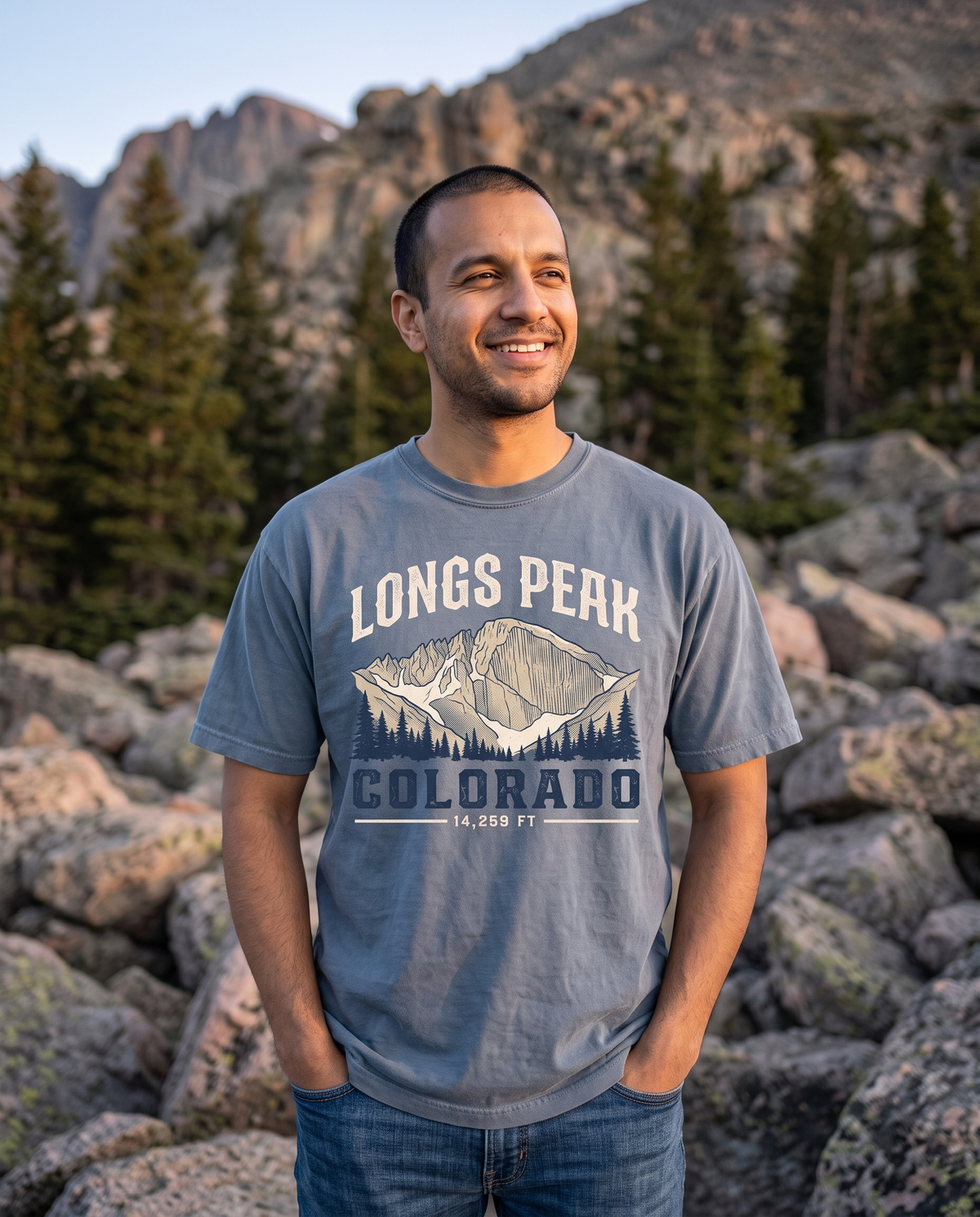A man stands on a granite boulder field below a rocky summit at dawn, hands in his pockets, grinning into the light.