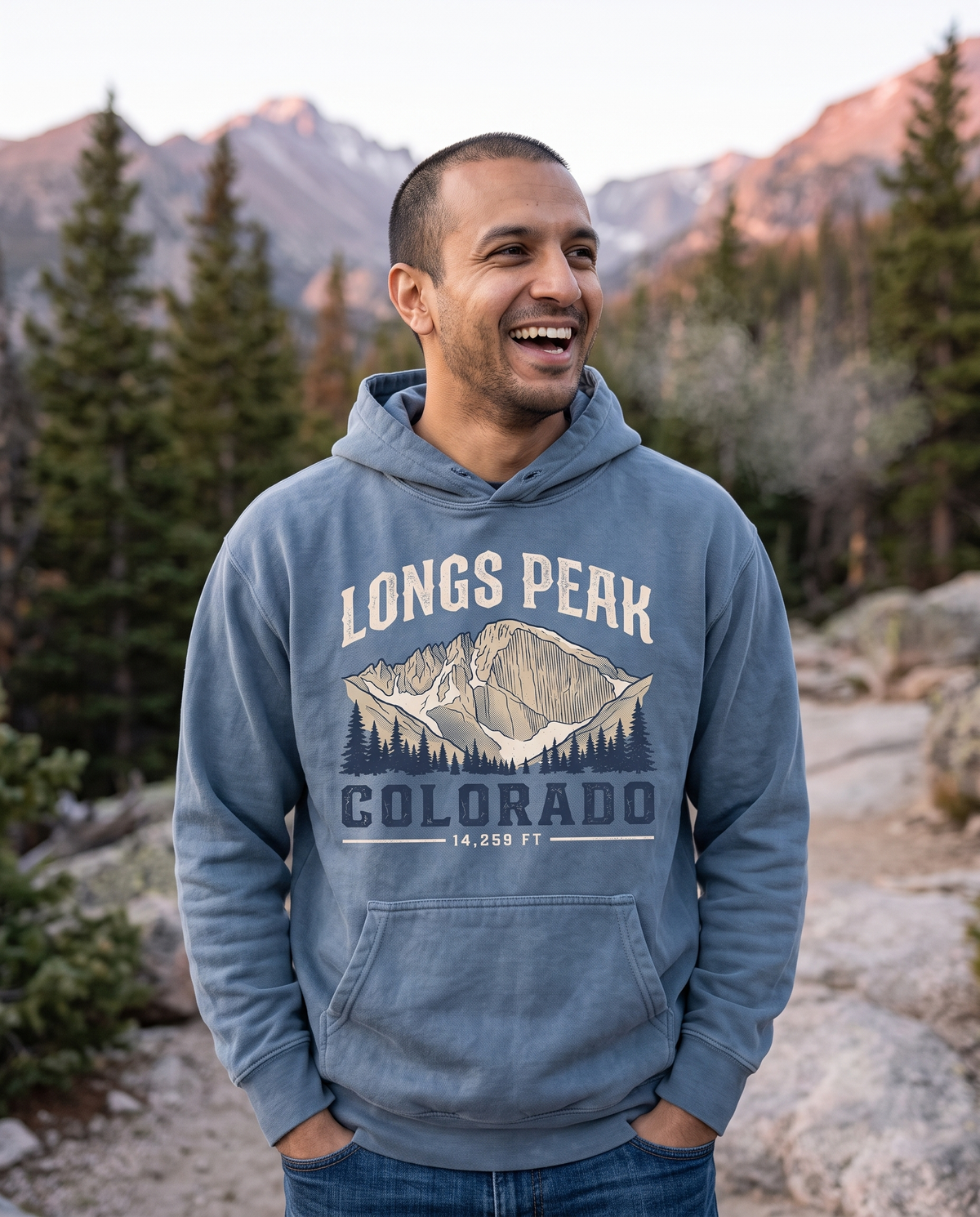 A man stands on a granite trail at sunrise with hands in his pockets, alpenglow on the peaks behind him.