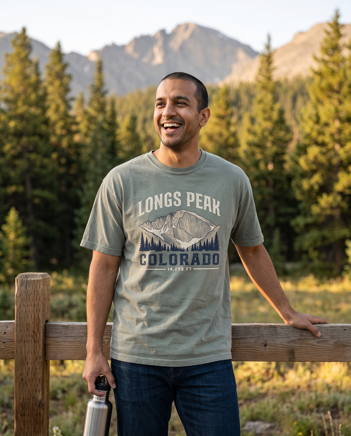 Man laughing at a trailhead fence in a Bay sage green Longs Peak Colorado 14,259 ft tee, lifestyle photograph