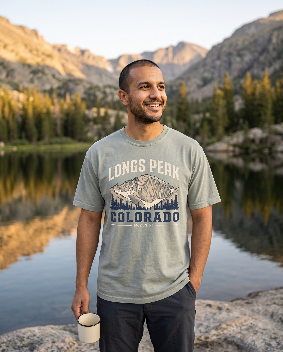 Man holding a camp mug beside an alpine lake at sunrise in a Bay sage green Longs Peak Colorado tee, lifestyle photograph