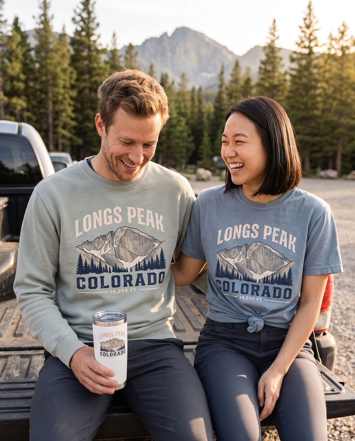 Two hikers laughing on a truck tailgate at a mountain trailhead wearing the Longs Peak Colorado 14,259 ft crewneck and tee, one holding a Longs Peak tumbler, lifestyle photograph