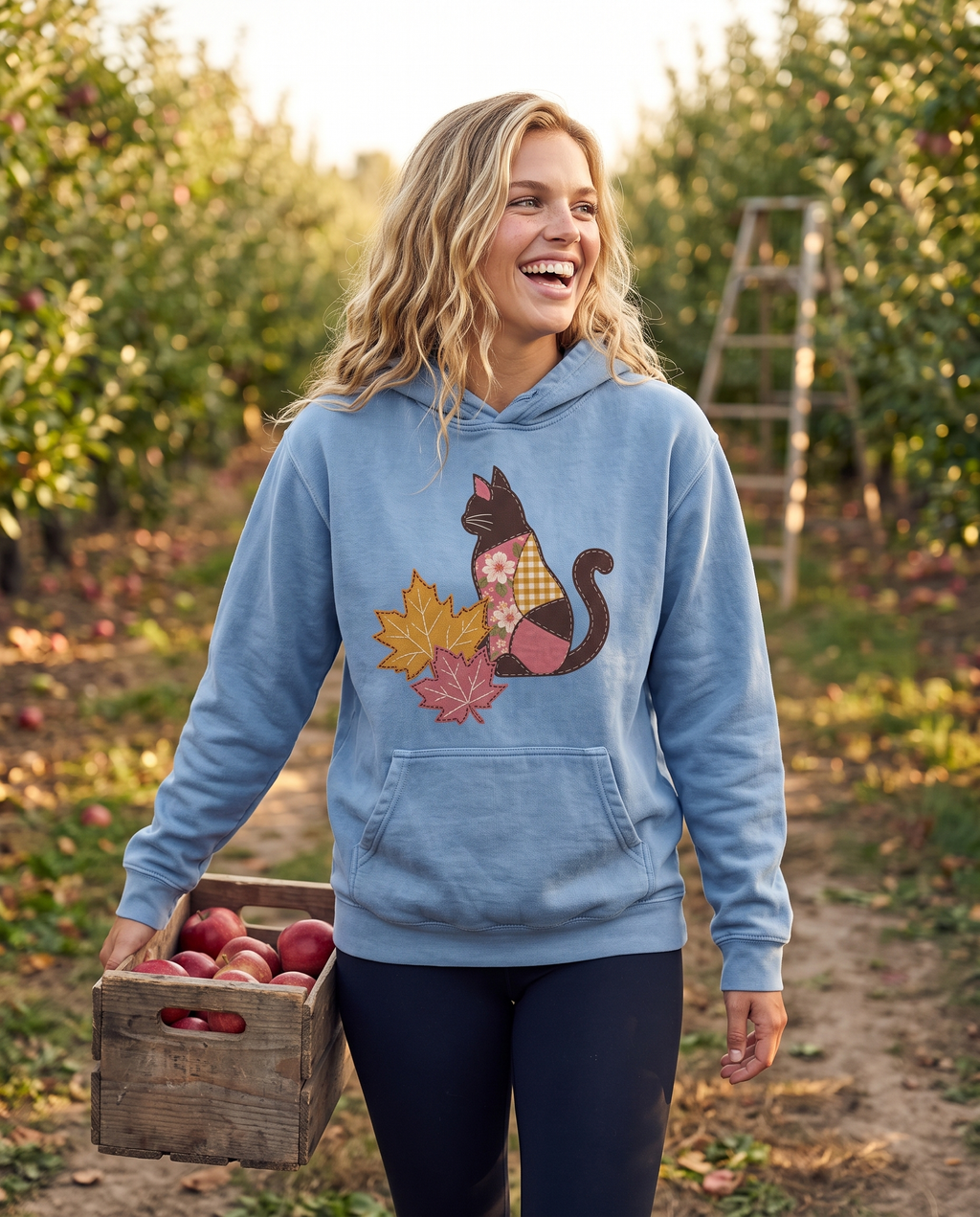 Woman carrying a crate of apples through an orchard in a blue hoodie printed with a patchwork black cat and two fall maple leaves, lifestyle photograph