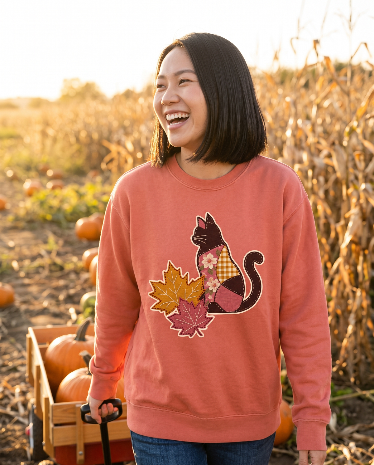 Woman laughing in a pumpkin patch wearing a Terracotta coral crewneck sweatshirt printed with a patchwork black cat and two fall maple leaves, lifestyle photograph