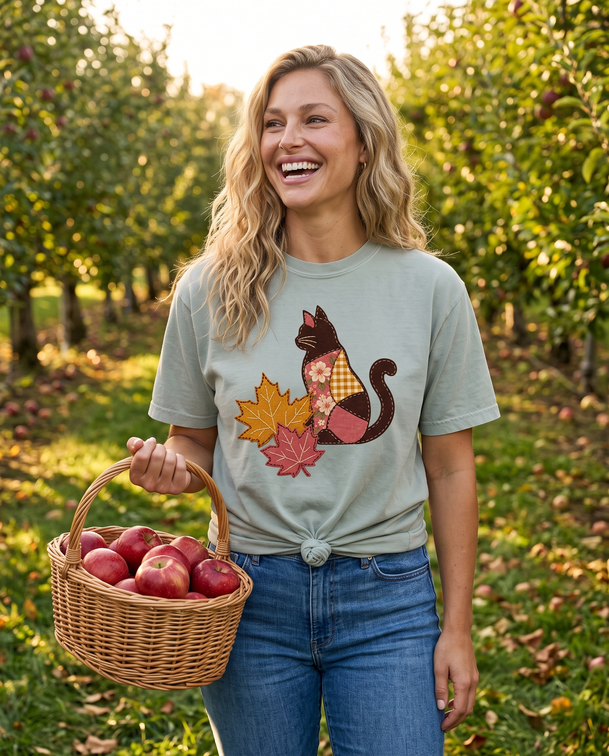 A laughing woman stands in a sunlit apple orchard holding a wicker basket of red apples, wearing a pale sage knotted tee and blue jeans.