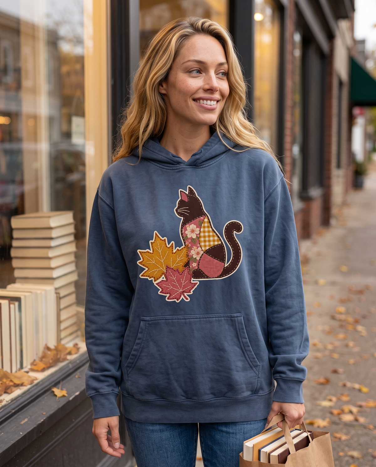 Woman outside a bookshop carrying a bag of books, wearing a Denim slate blue hoodie printed with a patchwork black cat and two fall maple leaves, lifestyle photograph