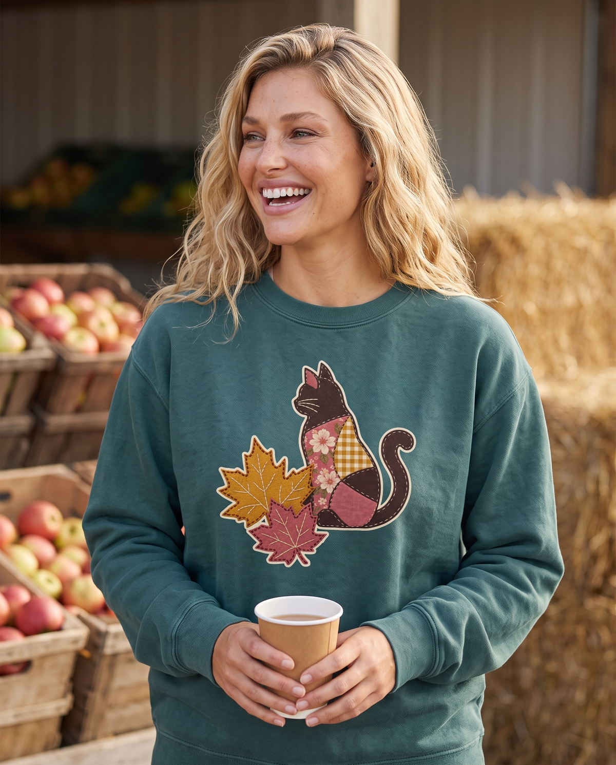 Woman at a farm stand holding a cup of cider, wearing a Blue Spruce green crewneck sweatshirt printed with a patchwork black cat and two fall maple leaves, lifestyle photograph