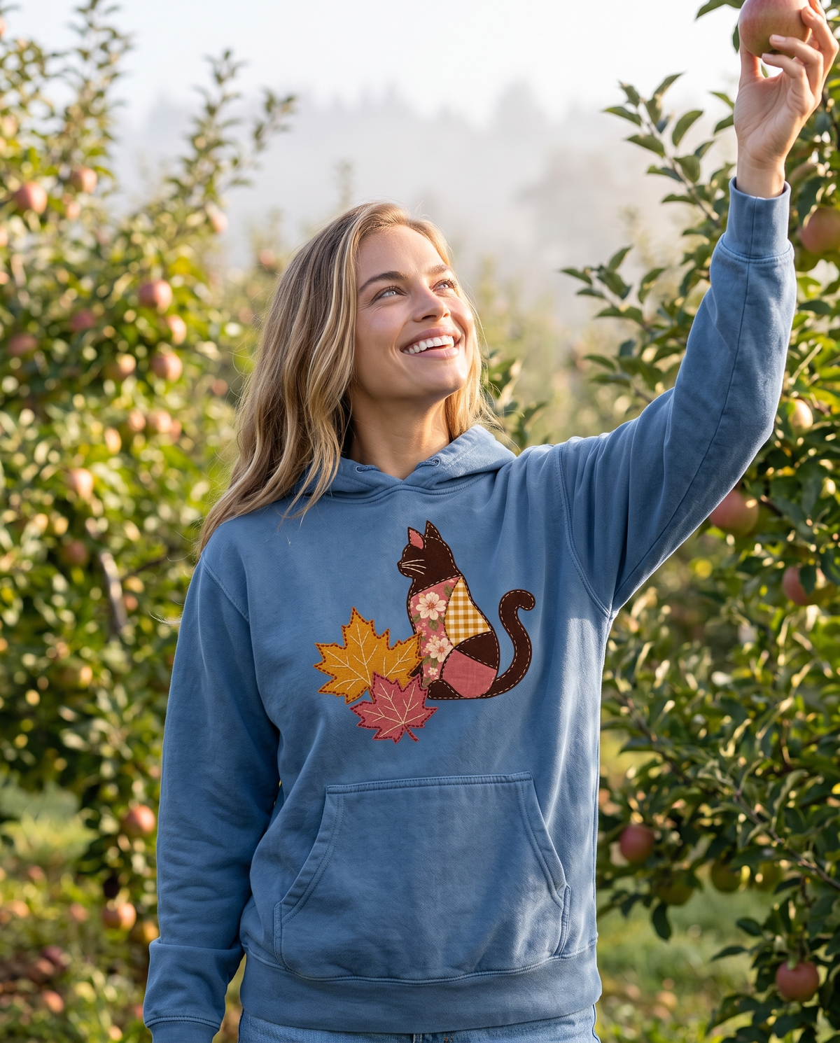 Woman picking an apple in an orchard wearing a Blue Jean hoodie printed with a patchwork black cat and two fall maple leaves, lifestyle photograph