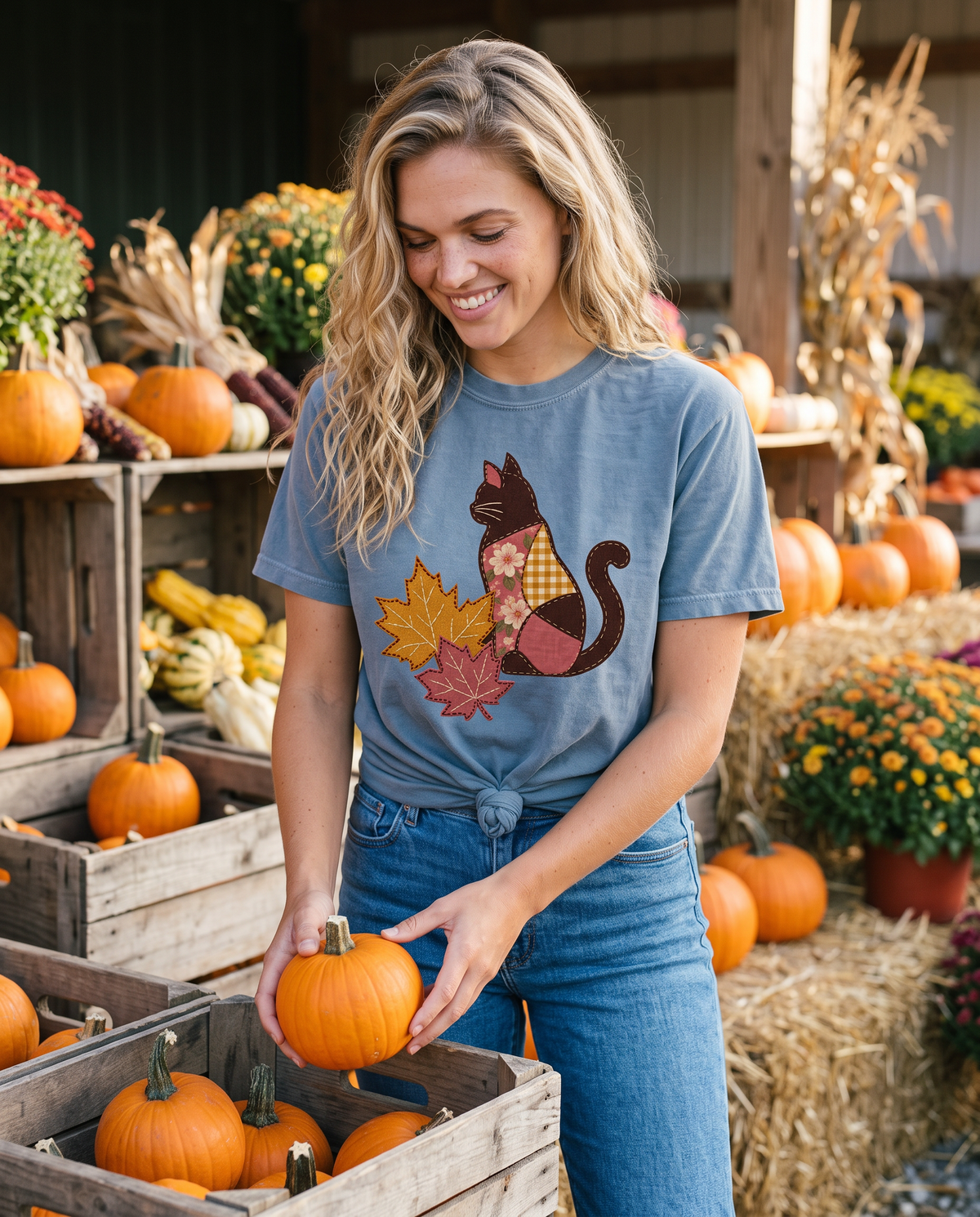 A smiling woman lifts a small orange pumpkin from a wooden crate at an autumn farm stand surrounded by gourds, mums and cornstalks.