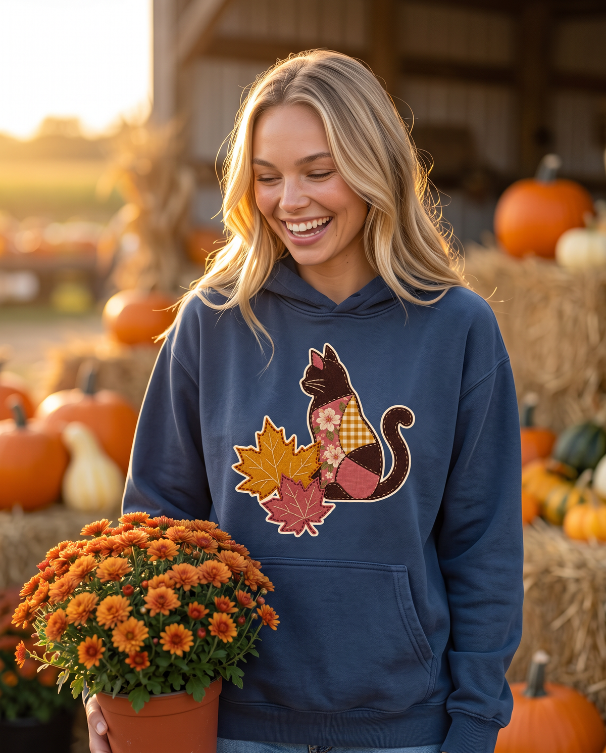 Woman at a farm stand carrying a pot of orange mums, wearing a True Navy hoodie printed with a patchwork black cat and two fall maple leaves, lifestyle photograph