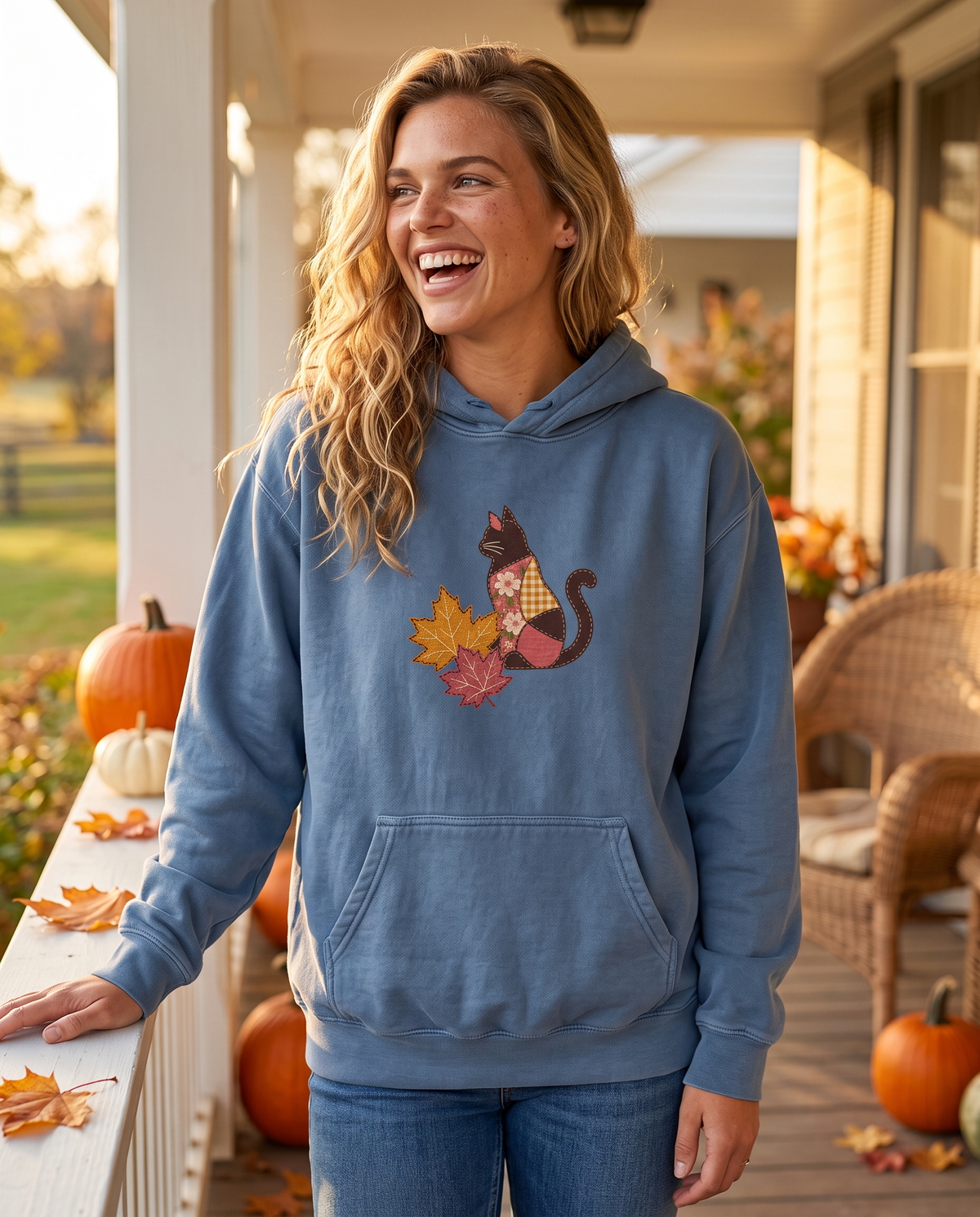 Woman laughing on a farmhouse porch wearing a Blue Jean hoodie printed with a patchwork black cat and two fall maple leaves, lifestyle photograph