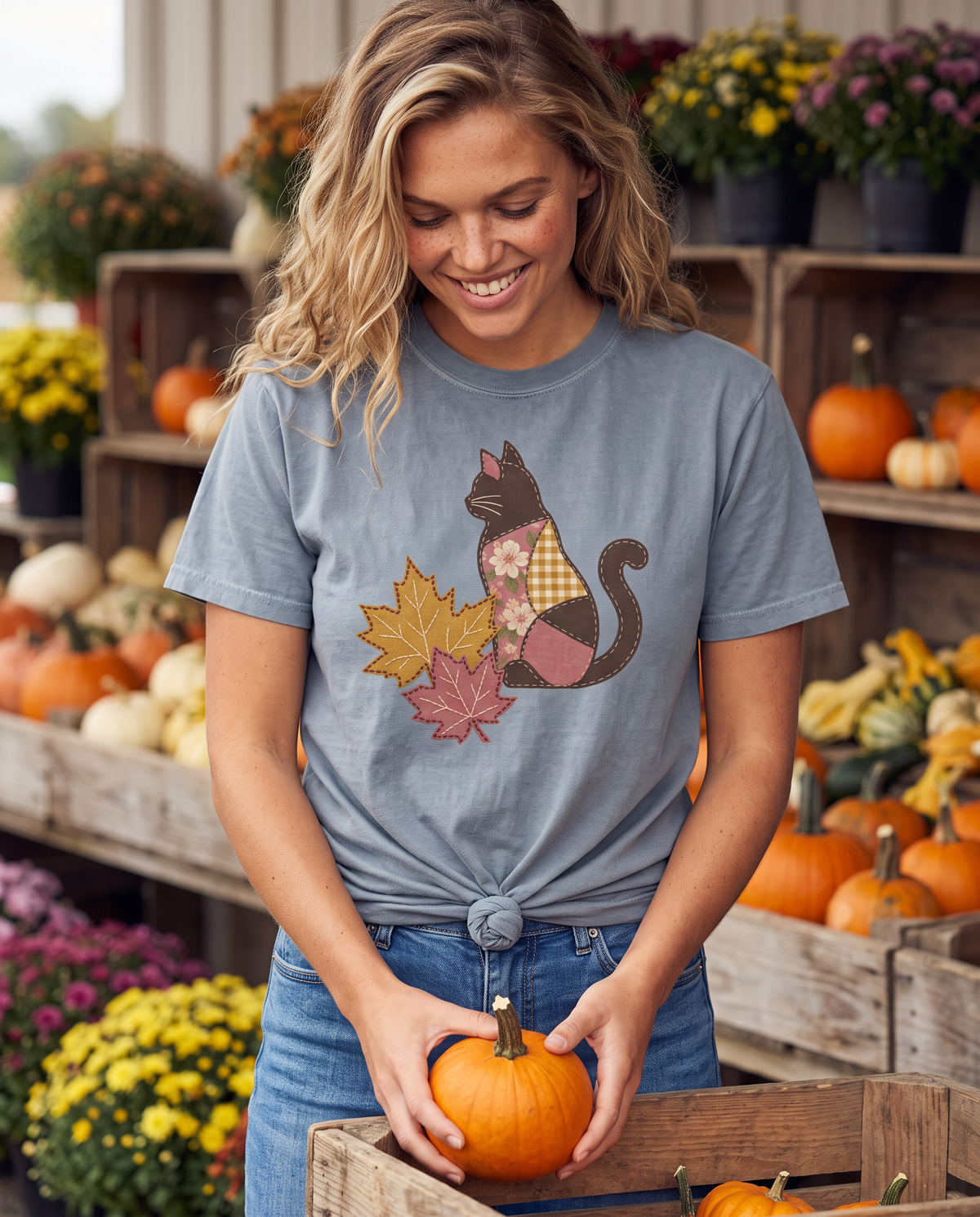 Smiling woman at a farm stand full of pumpkins and mums, holding a small pumpkin, wearing a blue-grey tee knotted at the hem with a patchwork black cat and fall leaf print.