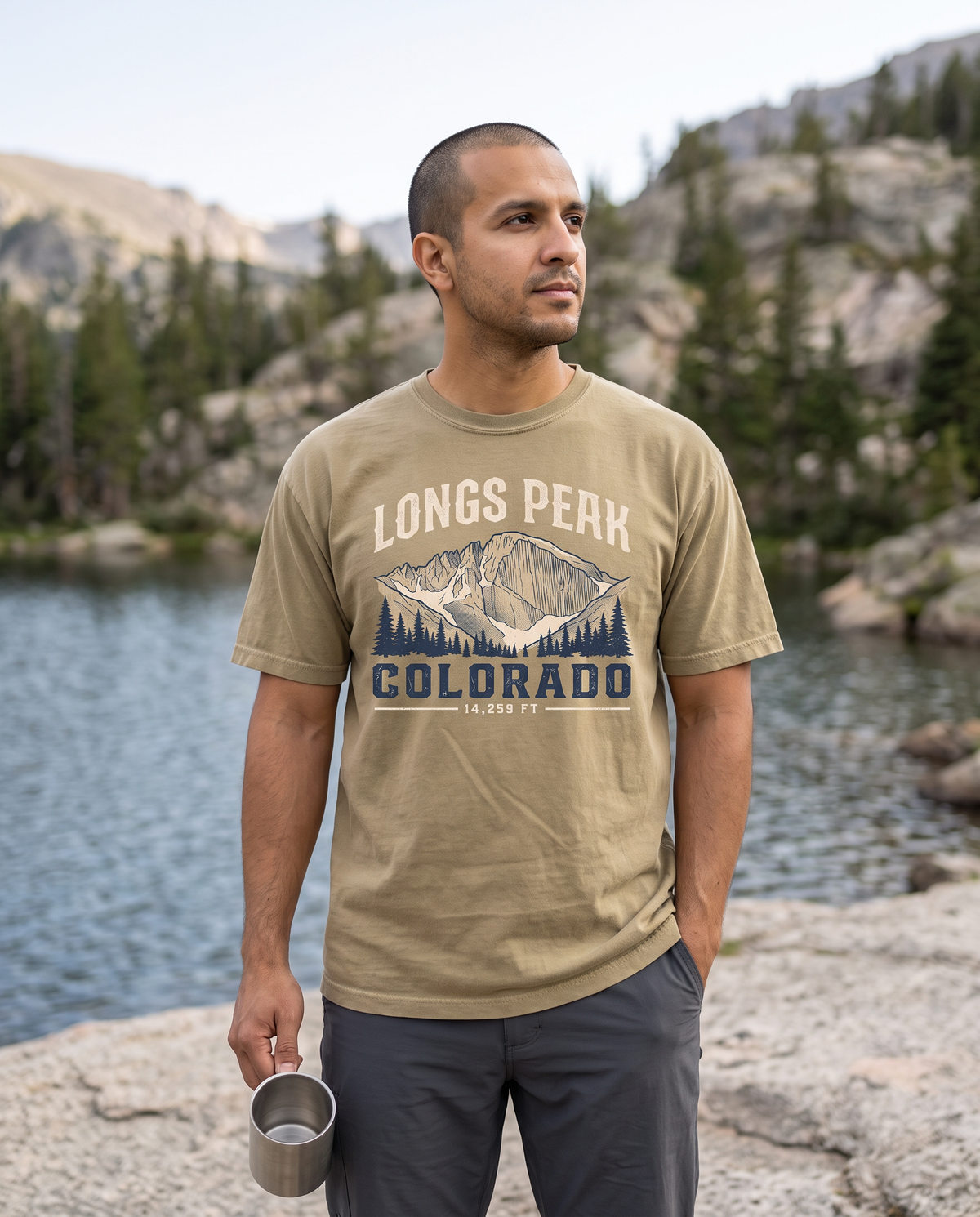 Man holding a camp mug beside an alpine lake with pines and granite, wearing a khaki tee printed LONGS PEAK COLORADO 14,259 FT with a mountain and pine graphic.
