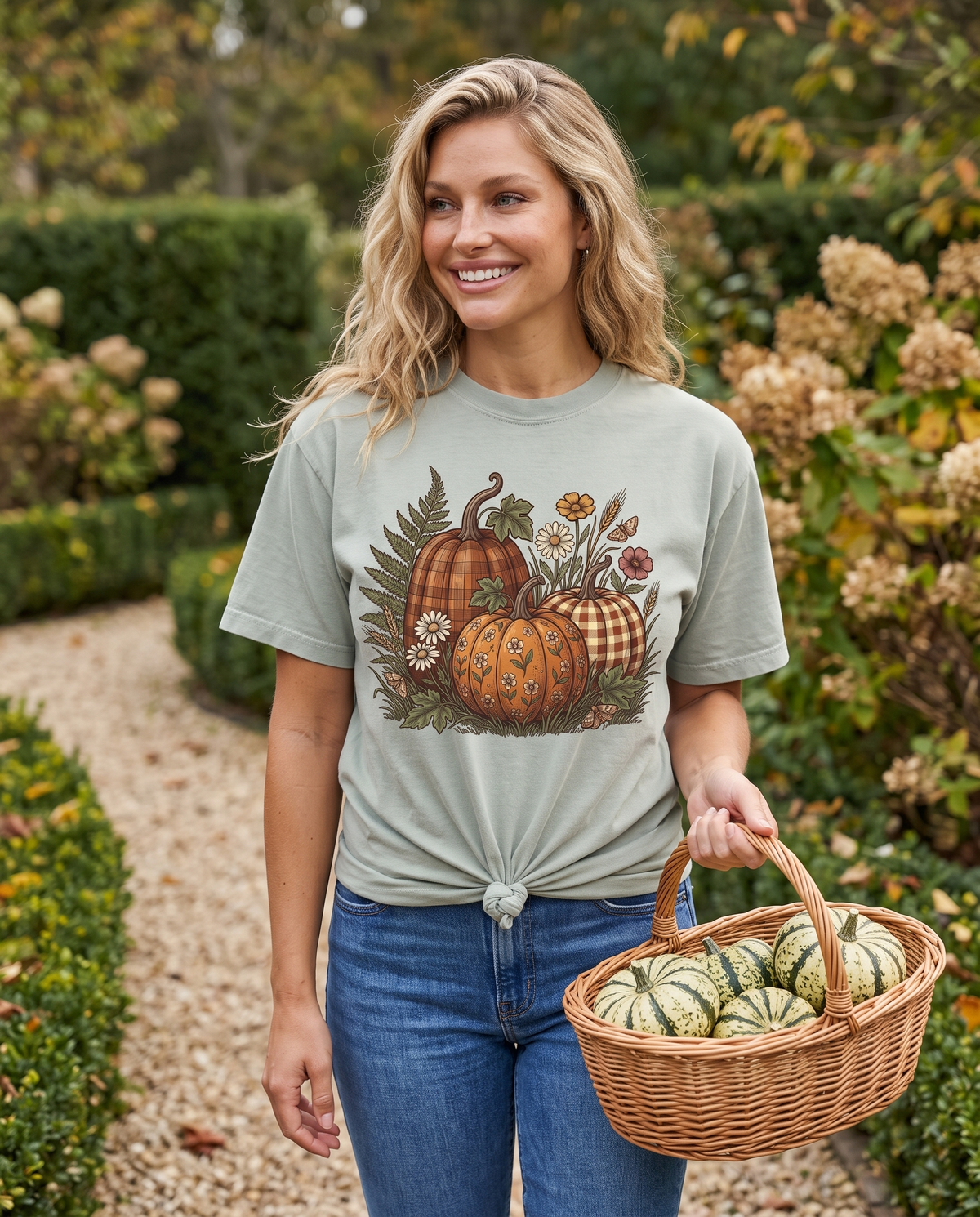 A woman walks a cottage garden path carrying a wicker basket of striped pumpkins, in a sage green tee knotted at the hem with a pumpkin, fern and daisy print, and blue jeans.