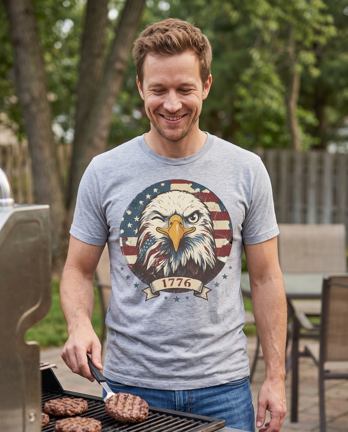 Smiling man grilling burgers on a backyard patio in a Sport Grey tee with the winking eagle and flag 1776 badge