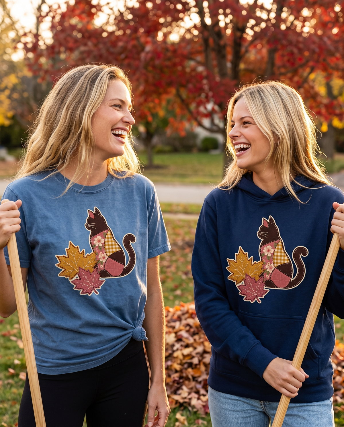 Two women laughing in an autumn yard of raked leaf piles, wearing a knotted Denim tee and a navy hoodie printed with a patchwork black cat and two fall leaves (A3-F14)