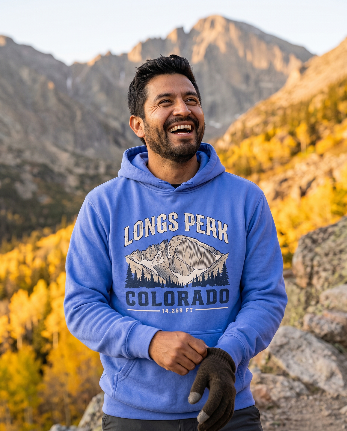 Laughing man in a Flo Blue hoodie with a mountain peak print pulling on a glove on a rocky trail with golden aspens and a granite peak behind