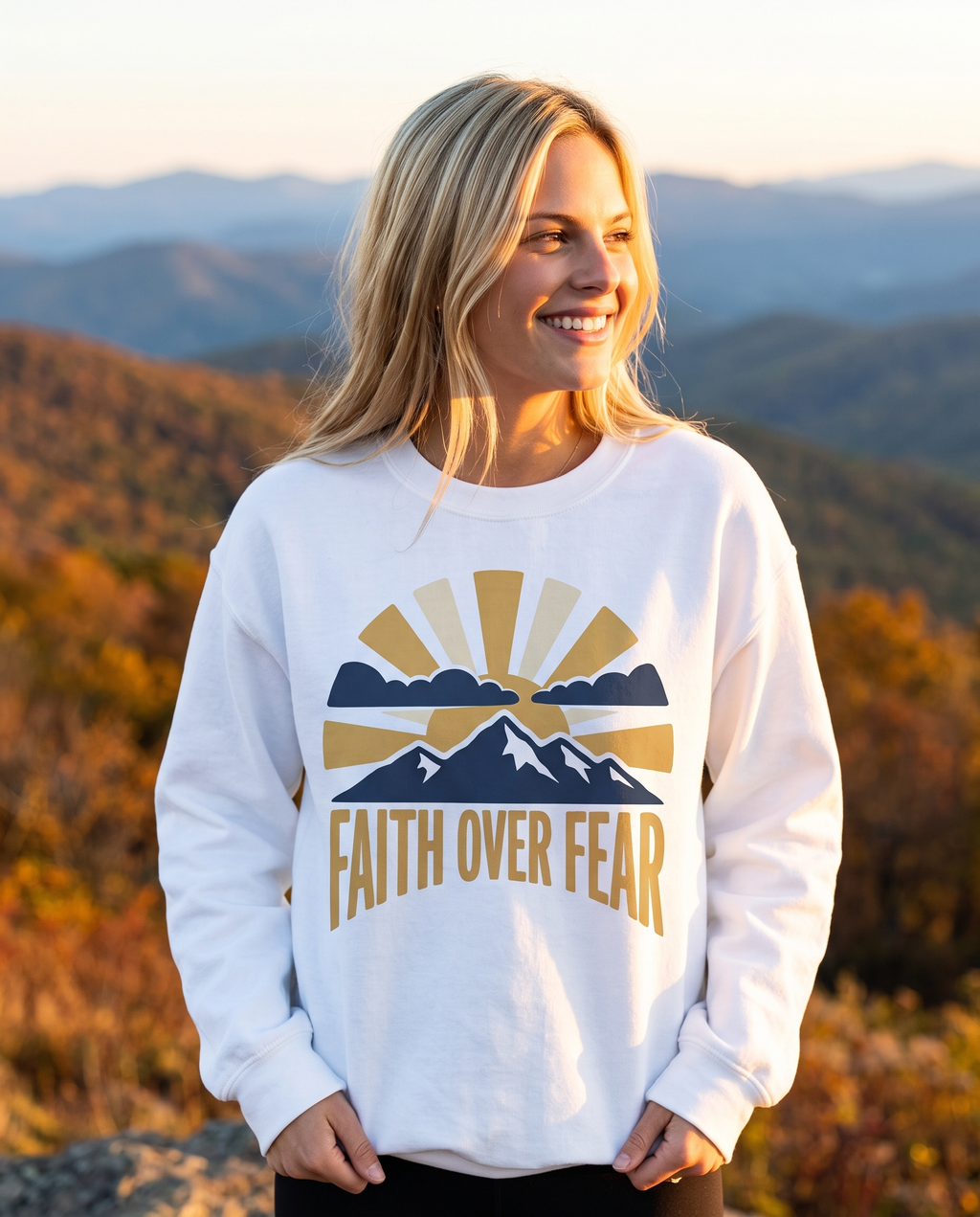 Smiling woman in a White sweatshirt with a sunrise and mountain print standing on an autumn ridge with layered blue mountains at sunset