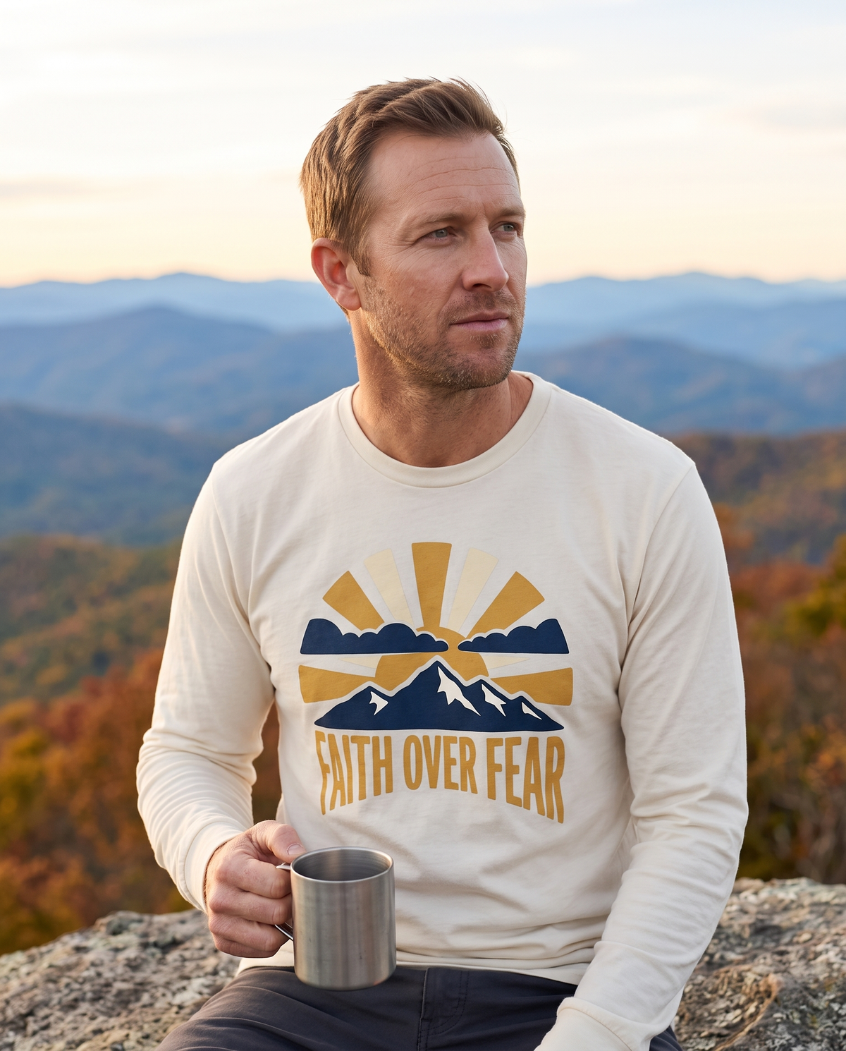 Man in an Ivory long-sleeve tee with a sunrise and mountain print sitting on a rock at a mountain overlook with a camp mug and autumn foliage