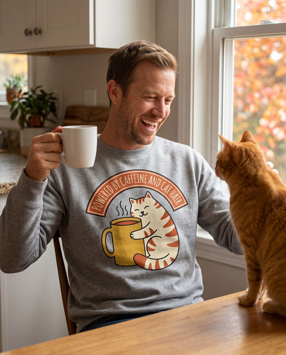 Laughing man in a Grey sweatshirt with a cat and coffee mug print holding a mug at a kitchen table beside an orange tabby cat and autumn window