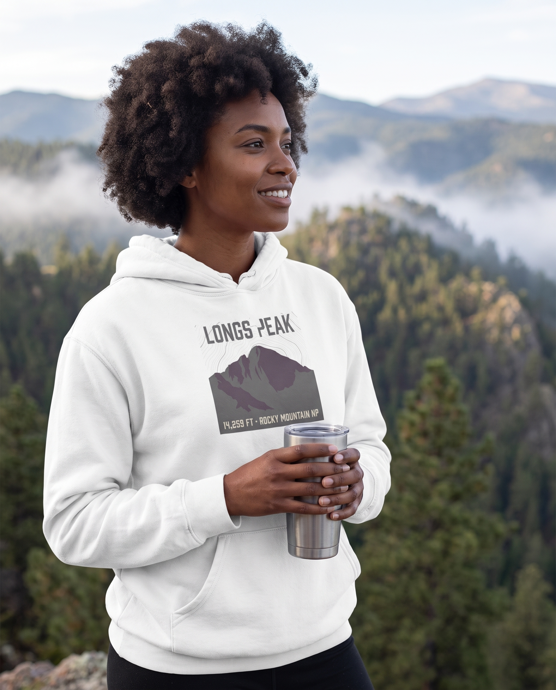 Woman at a misty Colorado mountain overlook wearing the Longs Peak 14,259 ft hoodie in White, topo summit badge design clearly visible on the chest