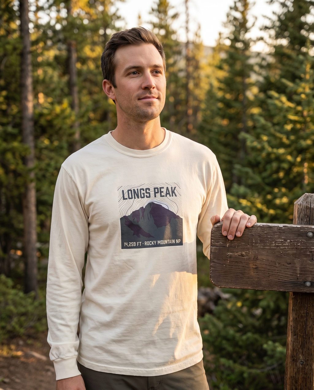 Man in a Colorado pine forest at golden hour wearing the Longs Peak 14,259 ft long sleeve tee in Ivory, topo summit badge design clearly visible on the chest