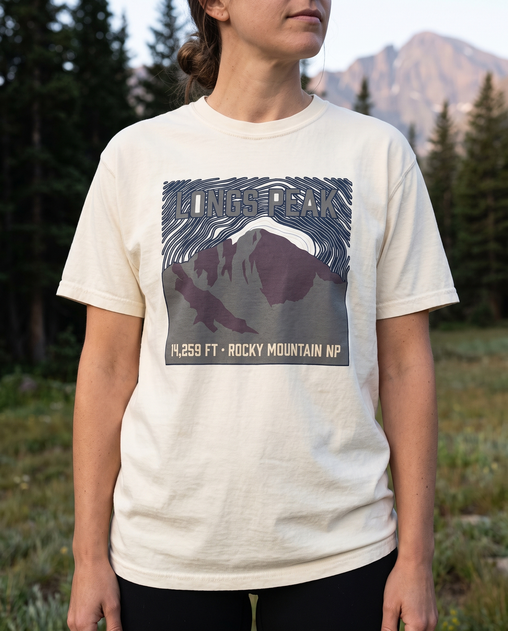 Woman at a Rocky Mountain National Park trailhead wearing the Longs Peak 14,259 ft tee in Ivory, navy topo badge design clearly visible on the chest