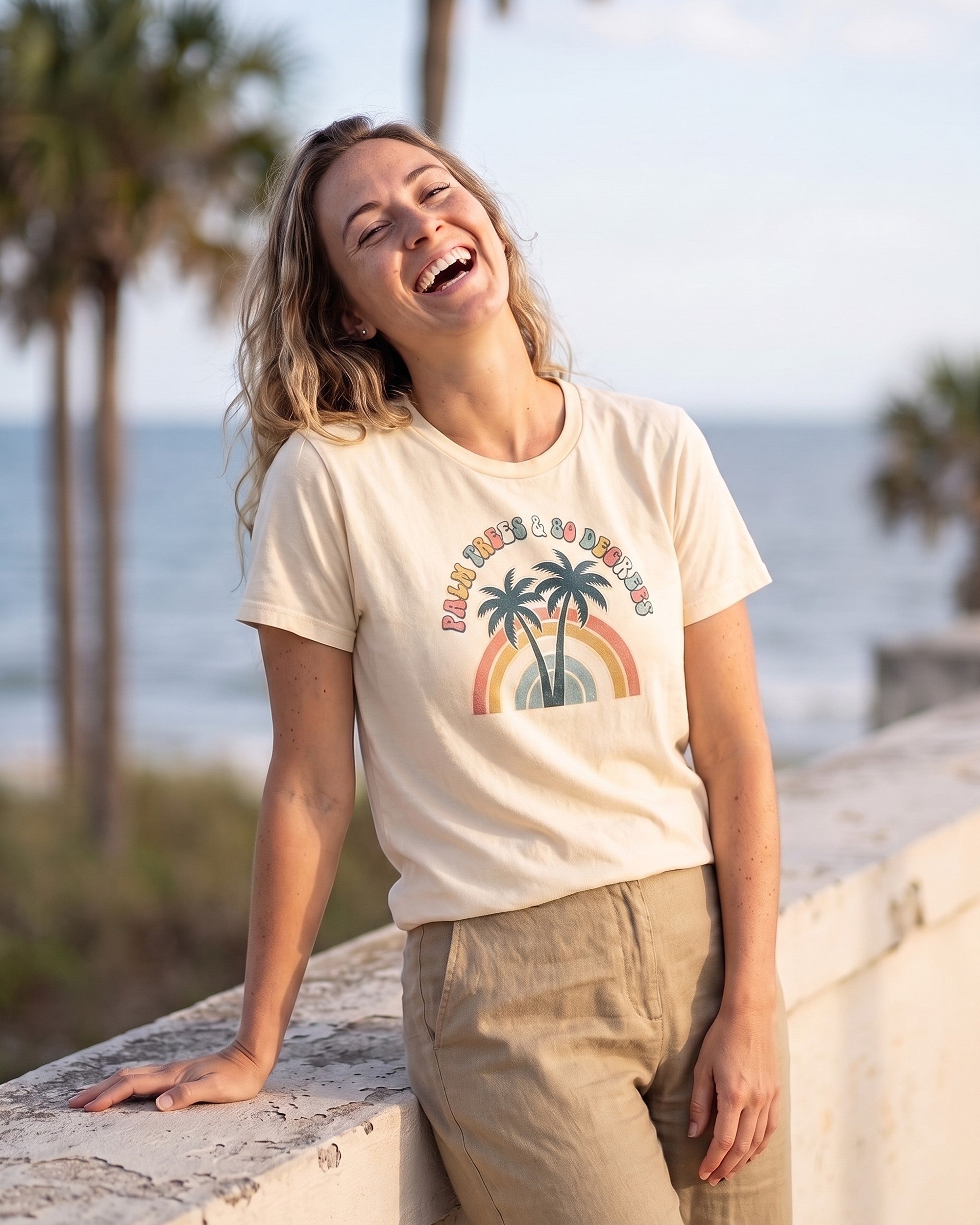 Woman on a palm-lined seafront wearing an ivory Palm Trees and 80 Degrees retro tee with two palms over a 1970s rainbow arch. AI generated model imagery, no real person photographed