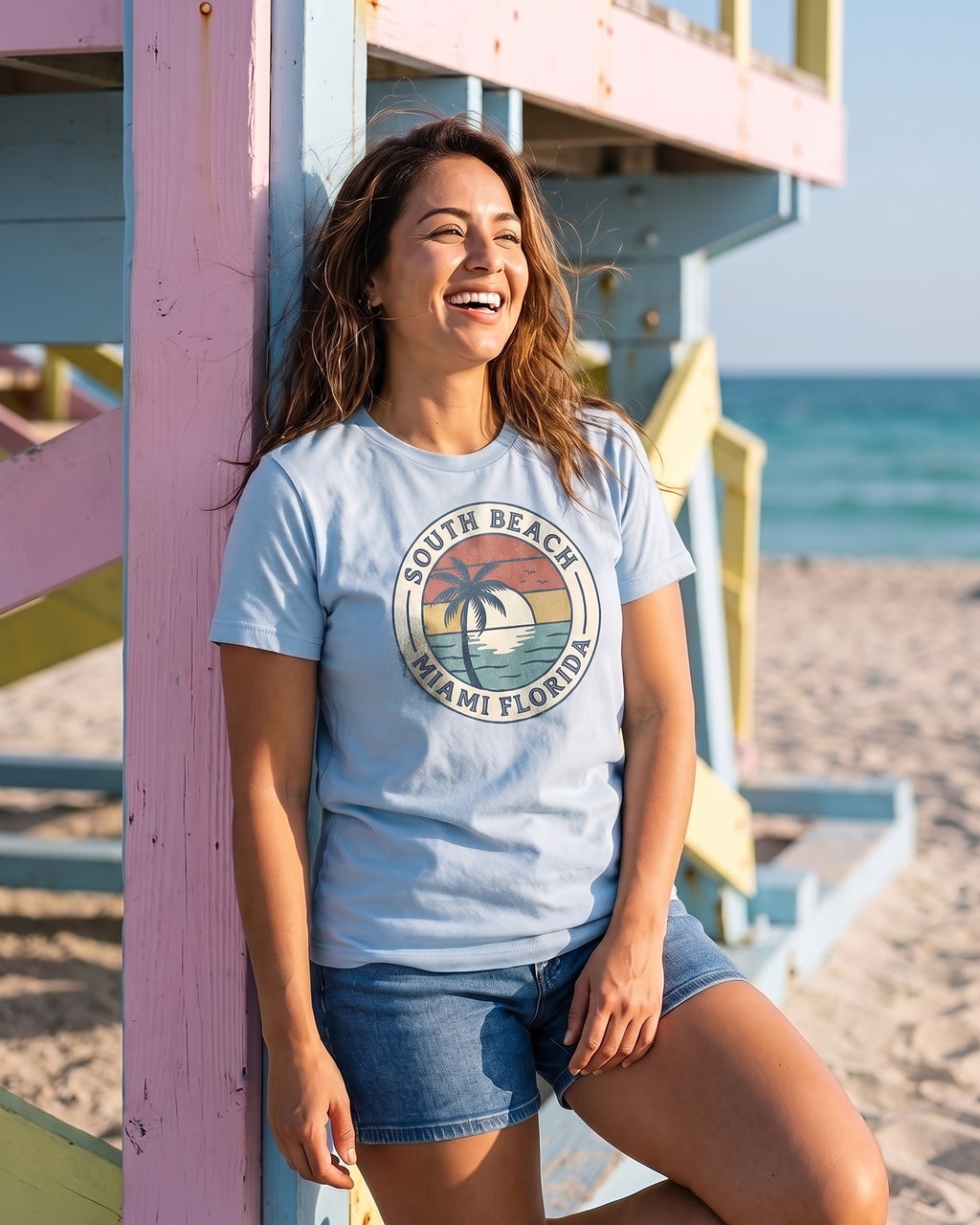 Woman leaning on a pastel South Beach lifeguard tower wearing a light blue South Beach Miami Florida vintage badge tee, the print following the folds of the cotton. AI generated model imagery, no real person photographed