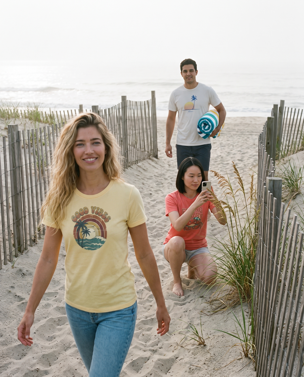 Morning dune path walk, the front friend in the Good Vibes retro beach tee in Butter, a friend photographing sea oats in the Feeling Crabby tee in Bright Salmon and a friend carrying a towel in the Retro Miami Palm tee in White