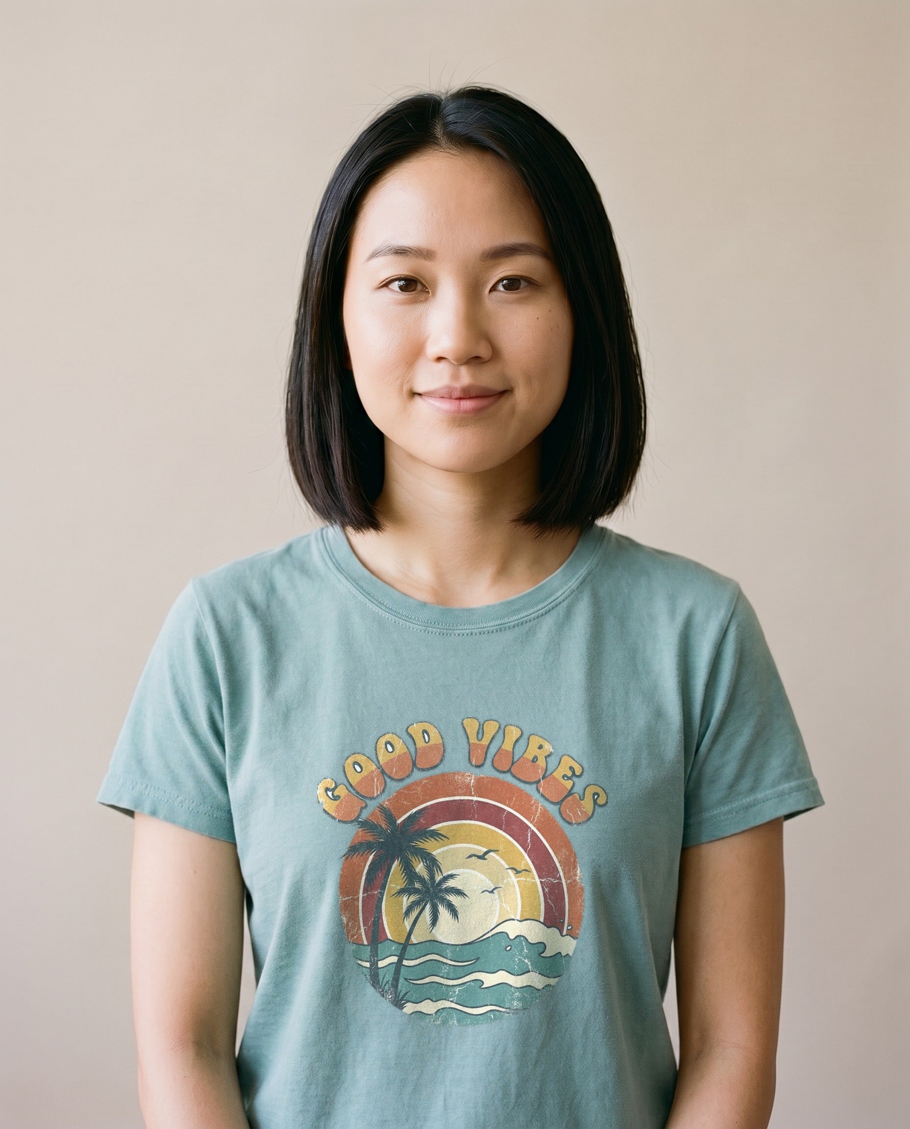 Woman wearing the Good Vibes retro beach tee in Bay sage green, arched Good Vibes lettering over palm and sunset